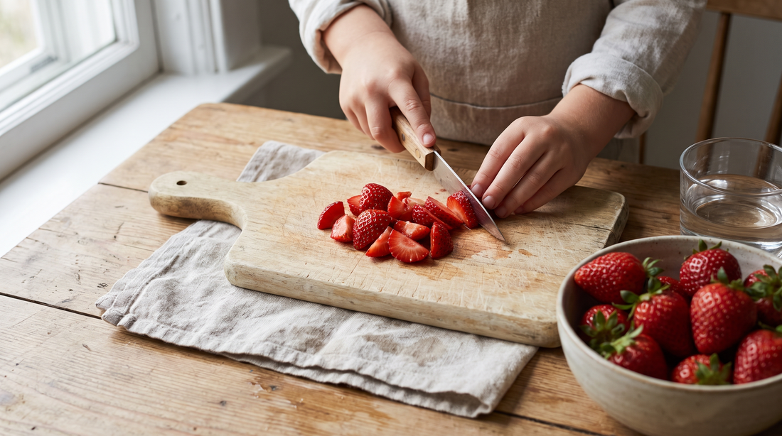 Child's hands slicing strawberries on a cutting board for kids at a wooden kitchen table