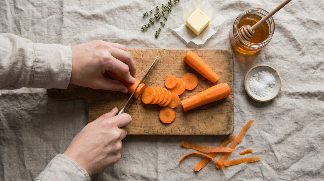 Hands slicing carrots into even coins on a wooden board before glazing them in a skillet