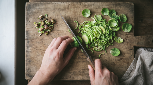 Hands slicing Brussels sprouts into thin ribbons on a wooden board for a weeknight side